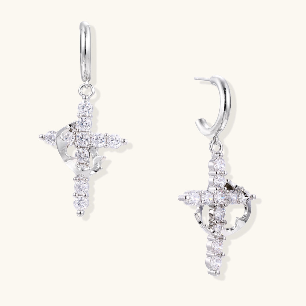 White gold-plated hoop earrings with sparkling zirconia cross charms dangling against cream background