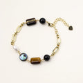 Gold chain bracelet with tiger's eye, obsidian, pearl, and iridescent abalone stones on cream background.