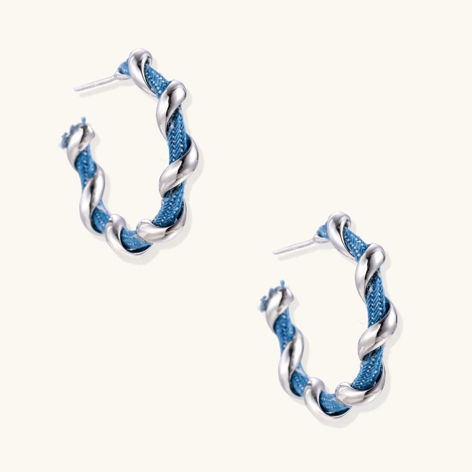 White gold-plated hoops intertwined with blue denim fabric in a twisted rope design on cream background.
