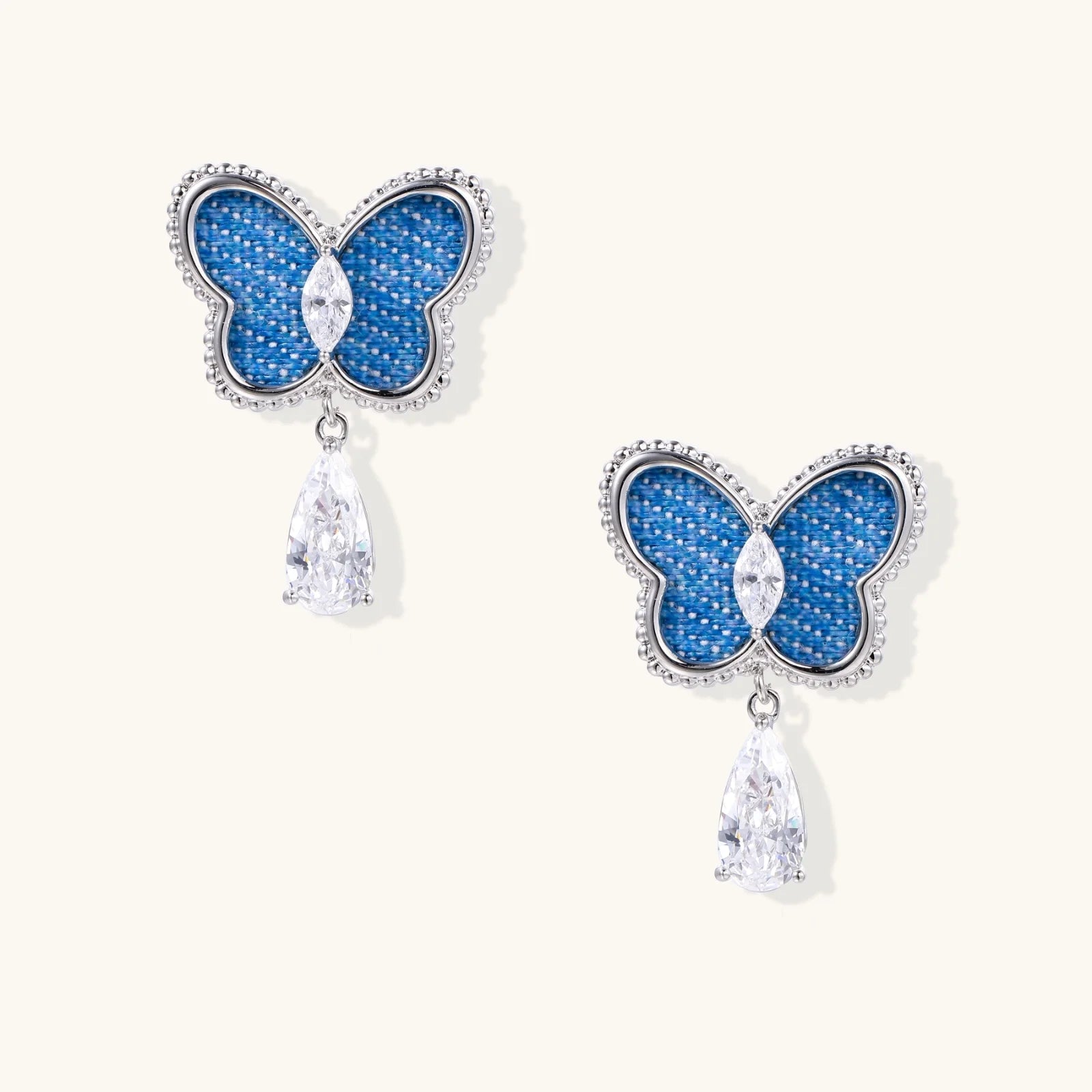 Blue denim butterfly earrings with silver frame and teardrop crystal dangles on cream background