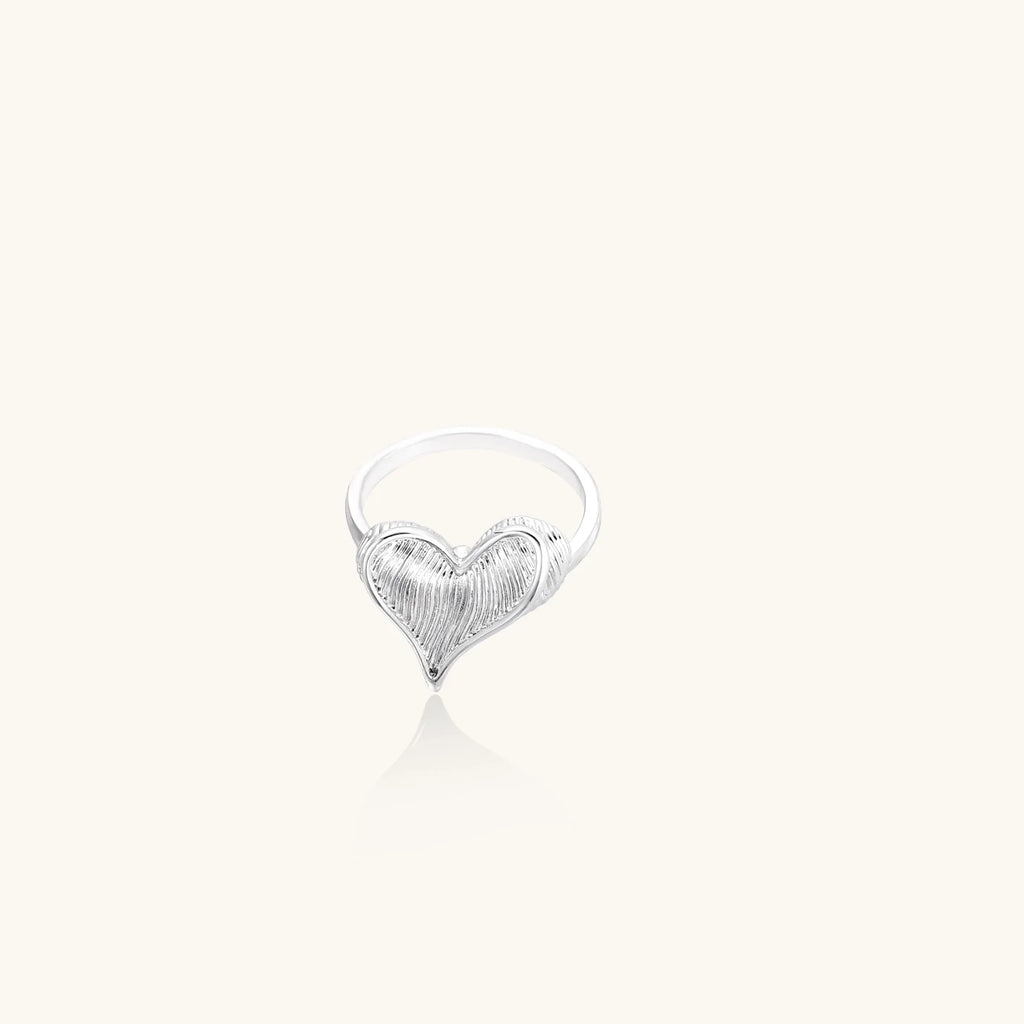 Silver ring with 3D heart formed by textured feather-like elements, white gold plated eco-brass on neutral background.