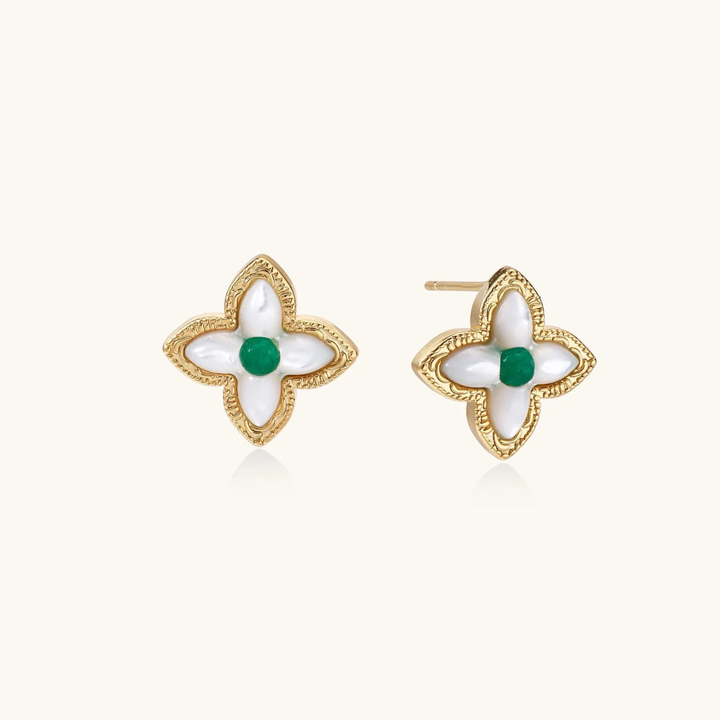 Gold quatrefoil stud earrings with white mother-of-pearl petals and emerald green gemstone center on beige background.