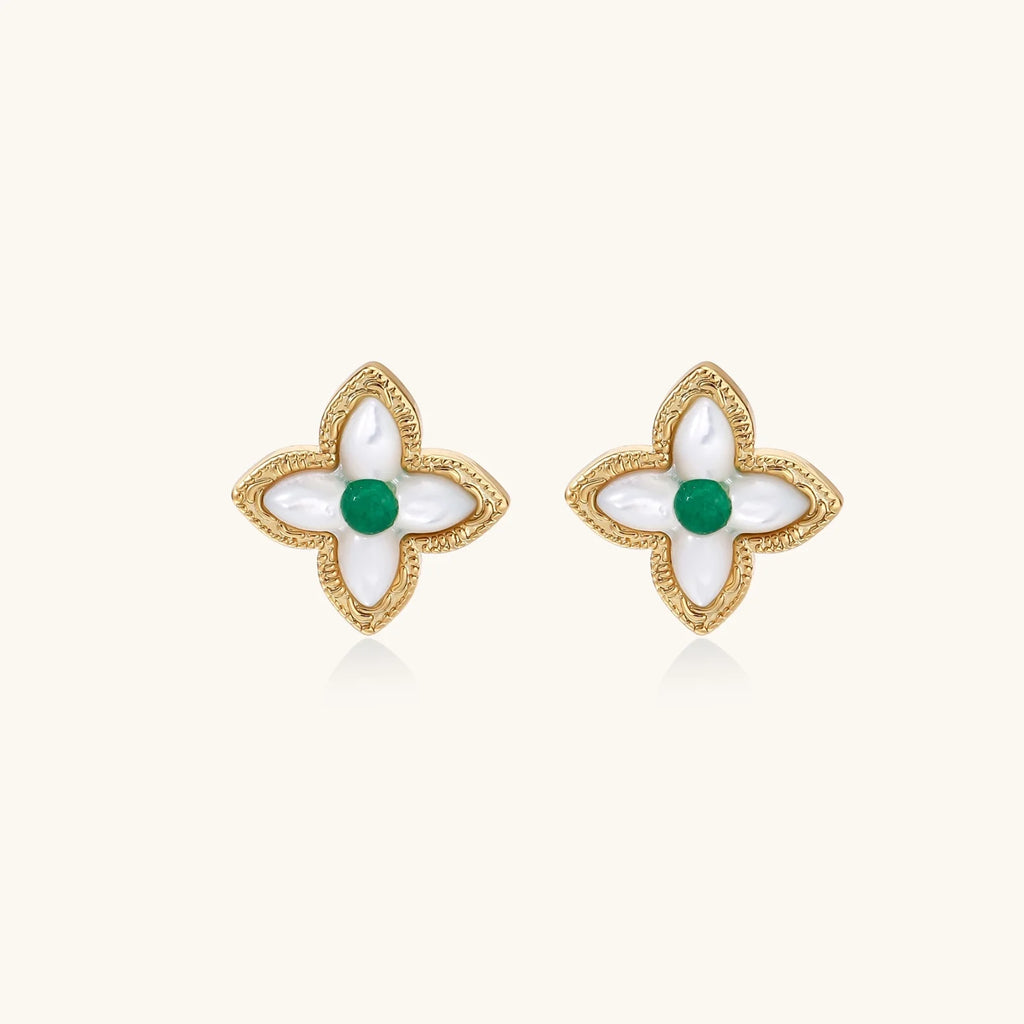Gold quatrefoil stud earrings with white mother-of-pearl petals and emerald green gemstone center on beige background.