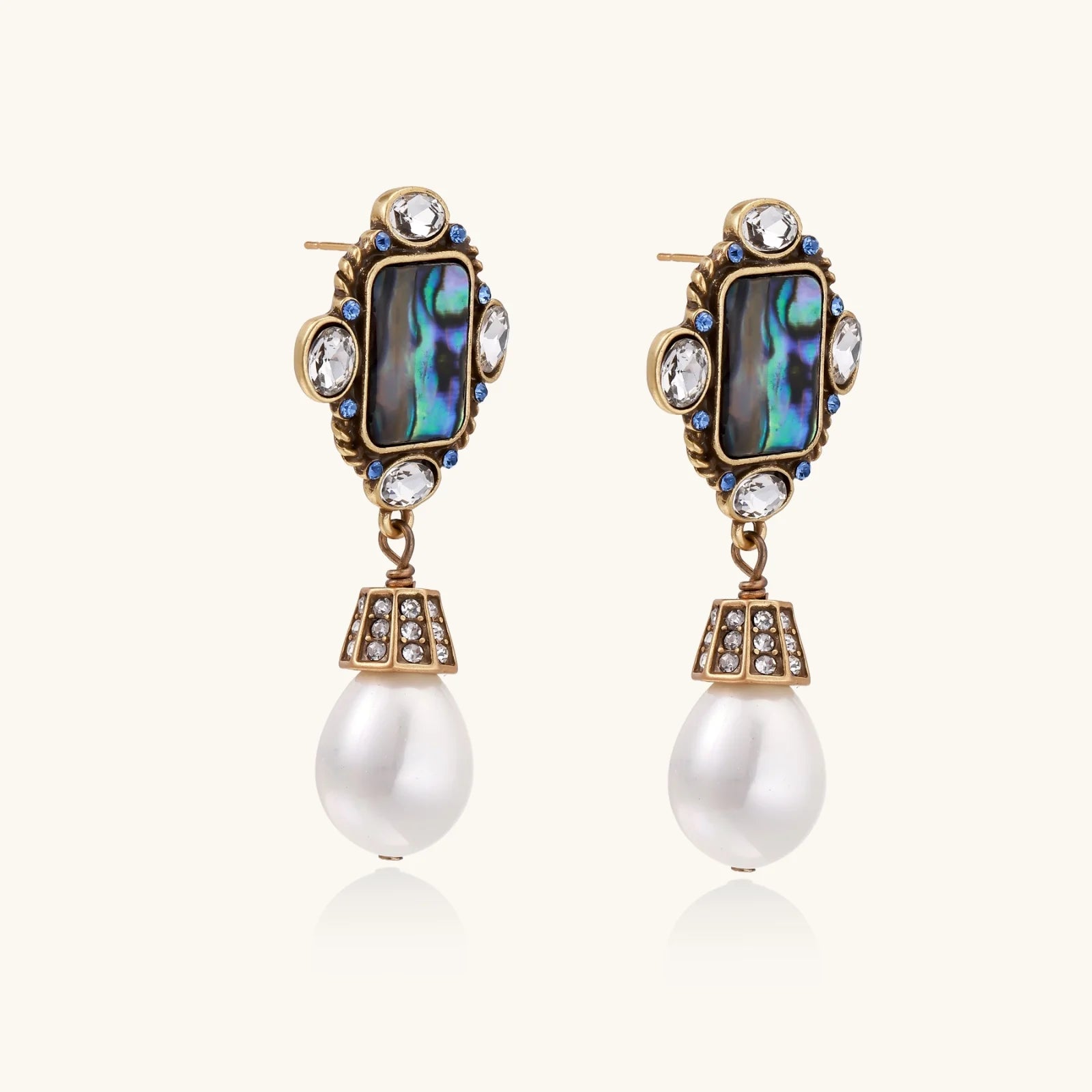 Ornate gold-plated earrings with iridescent abalone shell, crystals, blue stones, and white pearl drops on cream background.