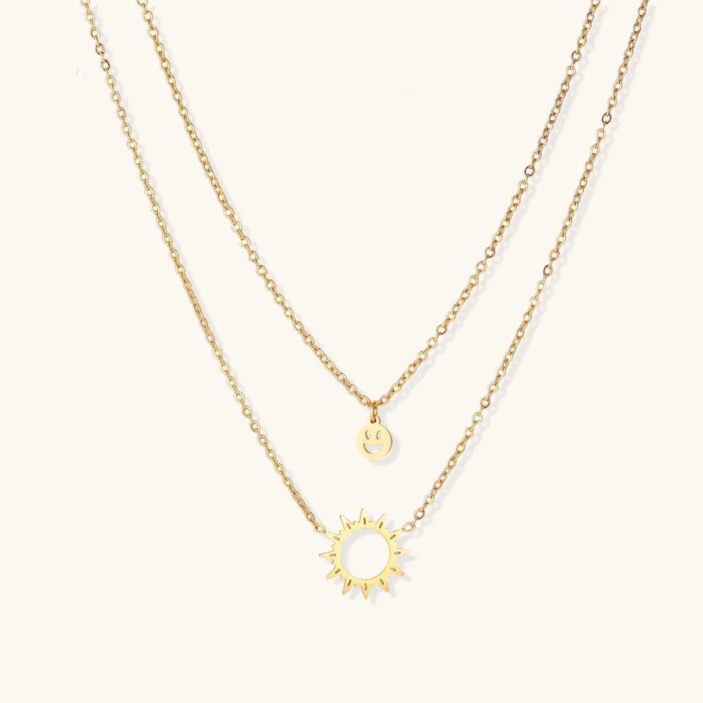 Gold layered necklace set with sunburst and smiley face pendants on delicate chains against white background.