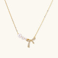 Delicate gold necklace with three white pearls and crystal-embellished bow pendant on cream background