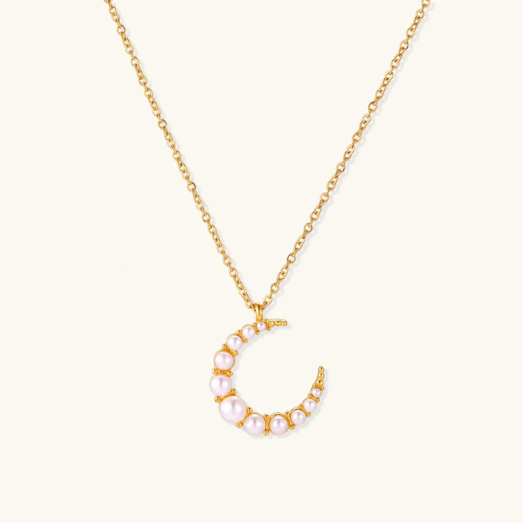 Gold crescent moon pendant with graduated white pearls on delicate chain against cream background