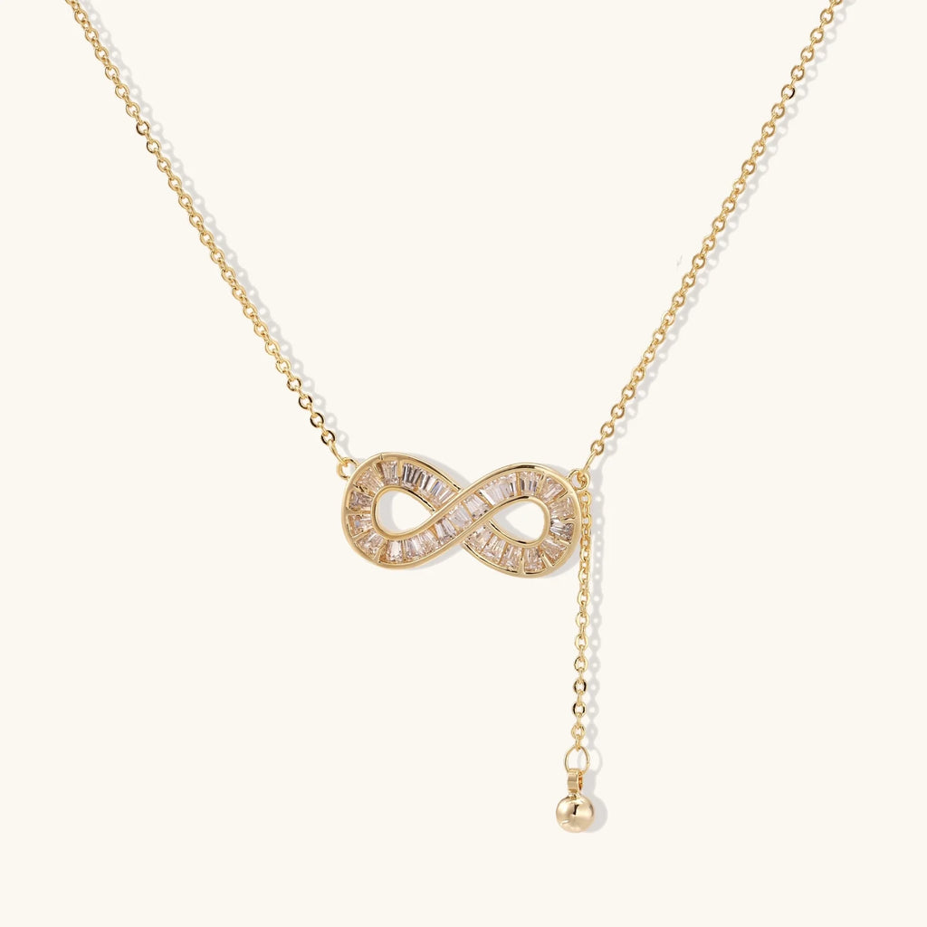 Gold infinity pendant with baguette crystals on adjustable chain against cream background.