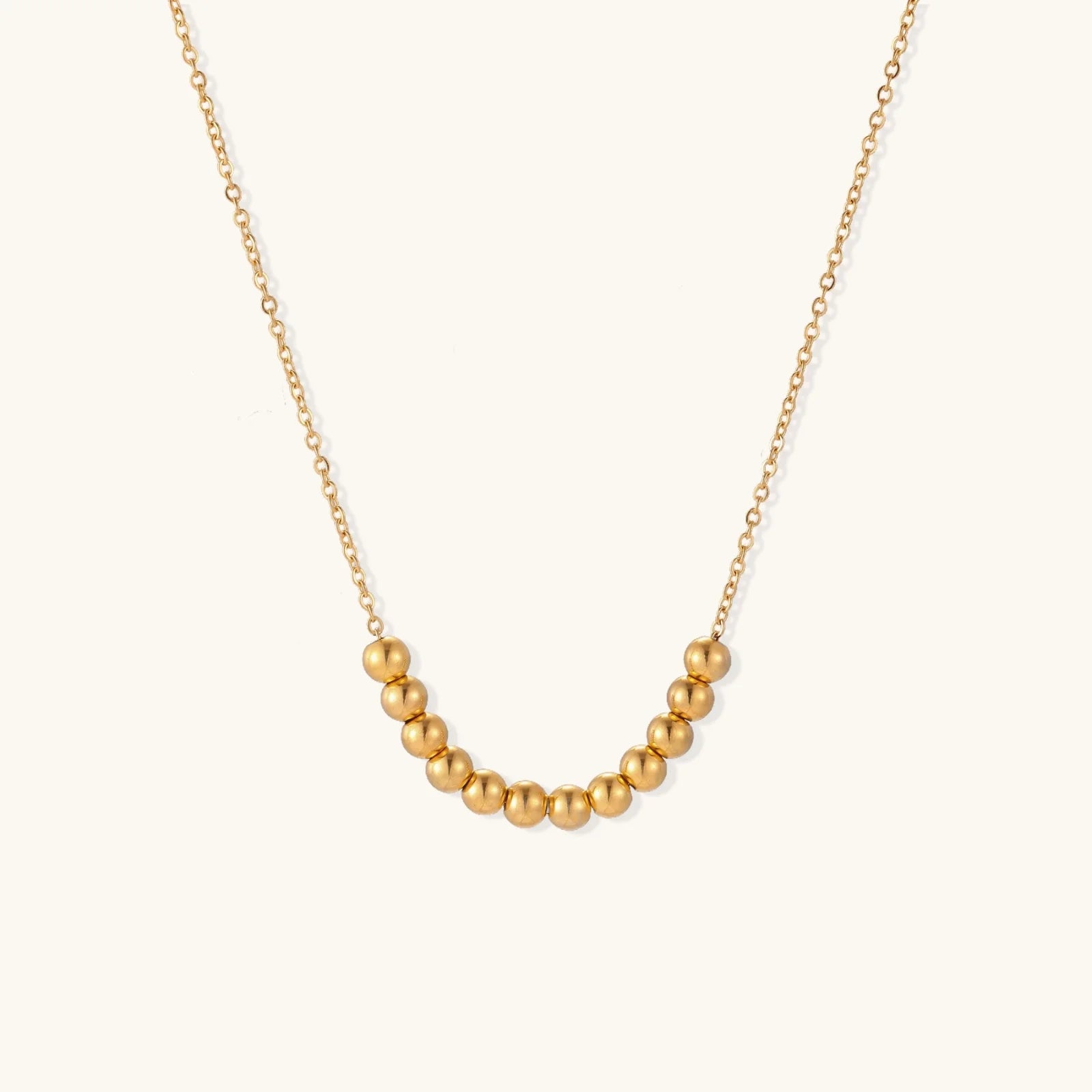 Delicate gold-plated necklace with graduated spherical beads forming gentle smile curve on beige background
